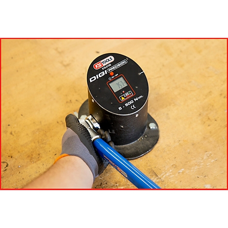 Eine Hand mit Handschuh bedient einen elektronischen Drehmomentschlüssel. Das Display zeigt einen Wert. Der Schlüssel ist auf einer Holzoberfläche platziert.